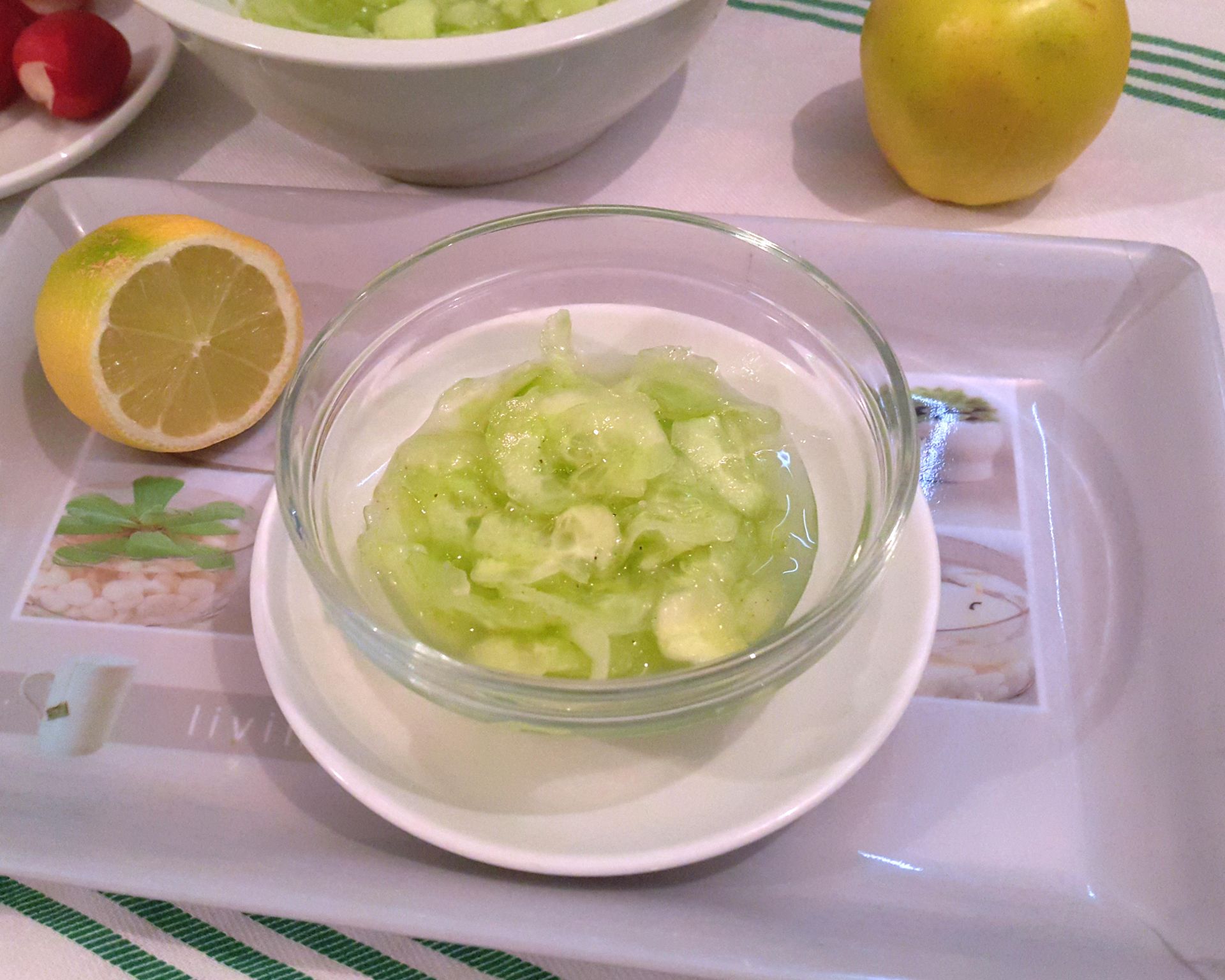 Šalátová uhorka nastrúhaná na tenké plátky v náleve v sklenenej miske na malom bielom tanieriku, v pozadí citrón