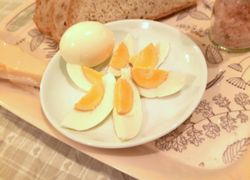 Krásne uvarené vajce na tvrdo prekrojené na mesiačiky na malom bielom tanieri na tácke, v pozadí chlieb a prílohy