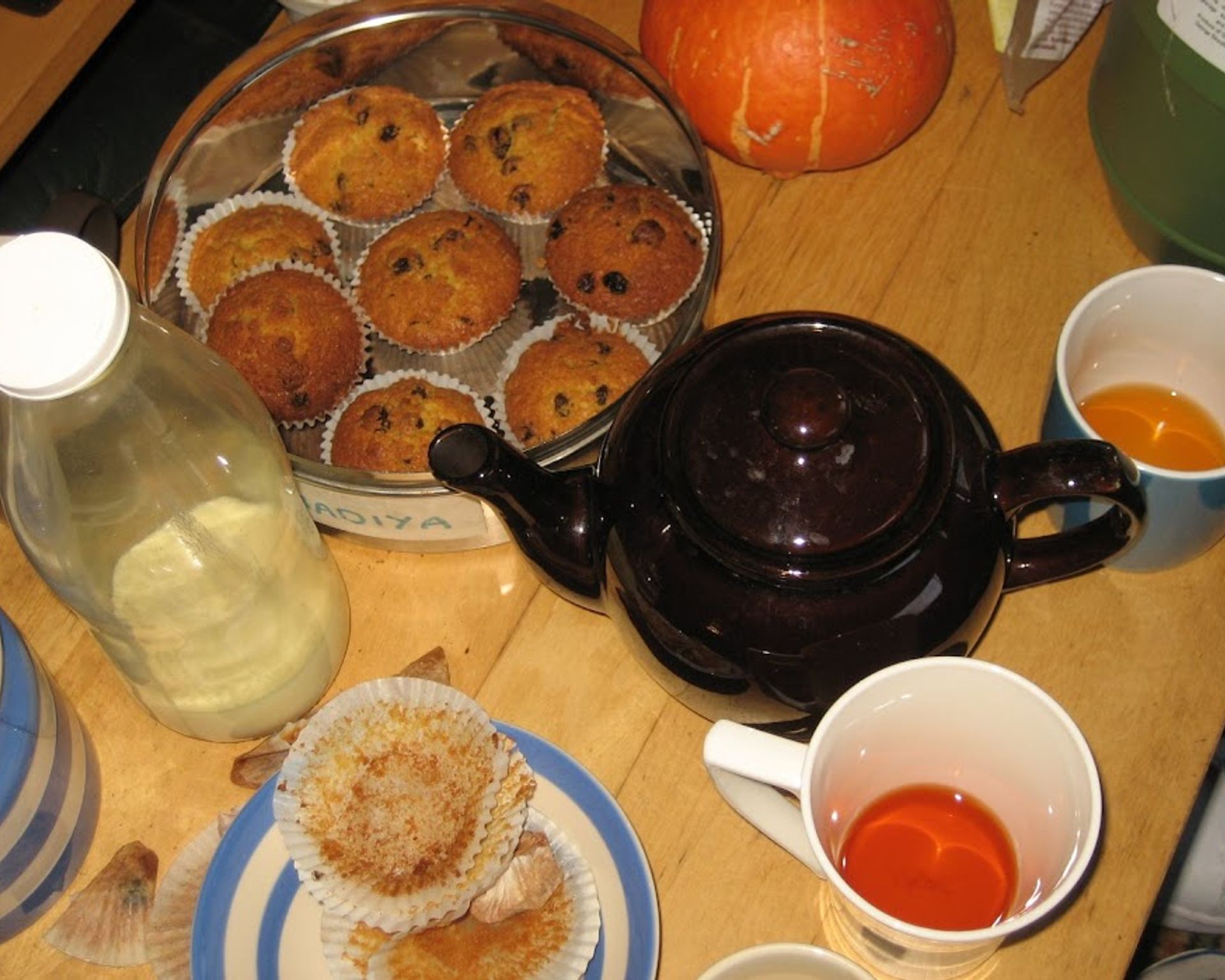 Muffiny v plechovej miske pri čajníku. Na stole sú aj čajové poháre, fľaša s mliekom a cukornička.