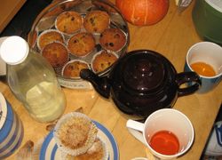 Muffiny v plechovej miske pri čajníku. Na stole sú aj čajové poháre, fľaša s mliekom a cukornička.
