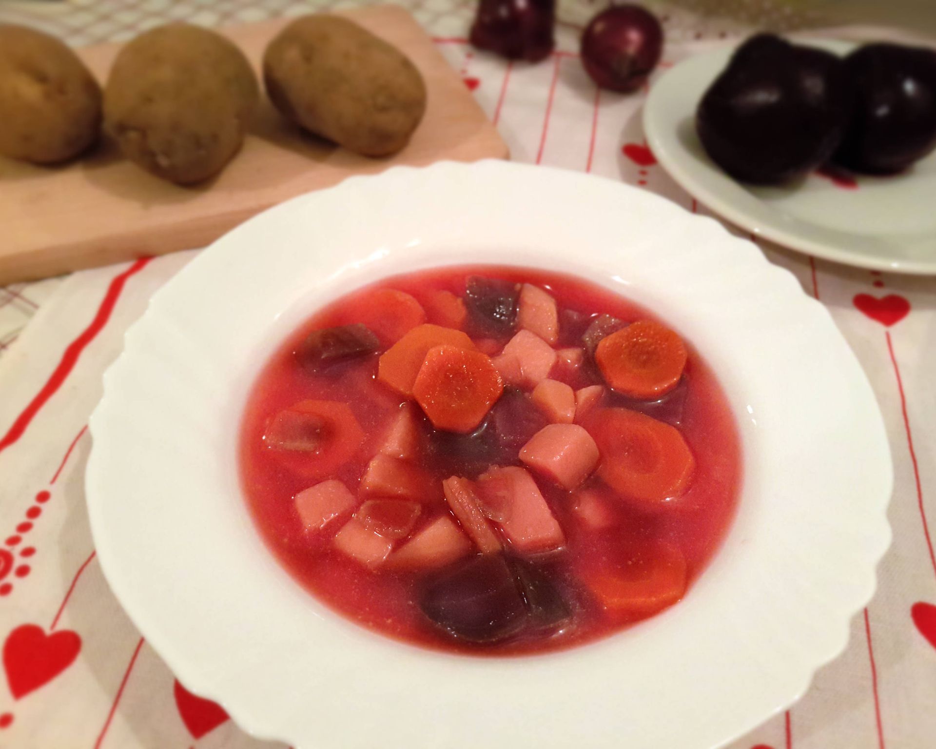 Na bielom tanieri naservírovaná polievka z červenej repy, mrkvy, zemiakov a cibule. V pozadí sú zemiaky surové a cvikla.