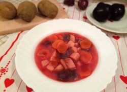 Na bielom tanieri naservírovaná polievka z červenej repy, mrkvy, zemiakov a cibule. V pozadí sú zemiaky surové a cvikla.
