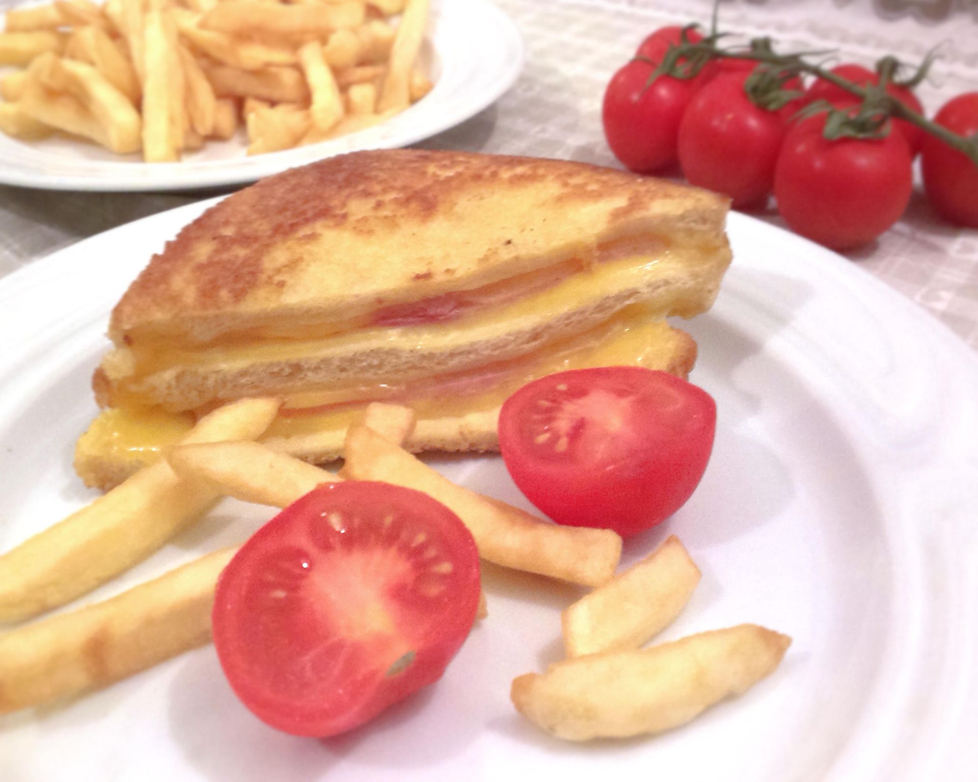 Sendvič plnený, v ňom šunka a syr, na tanieri aj hranolky a cherry paradajky, v pozadí tiež