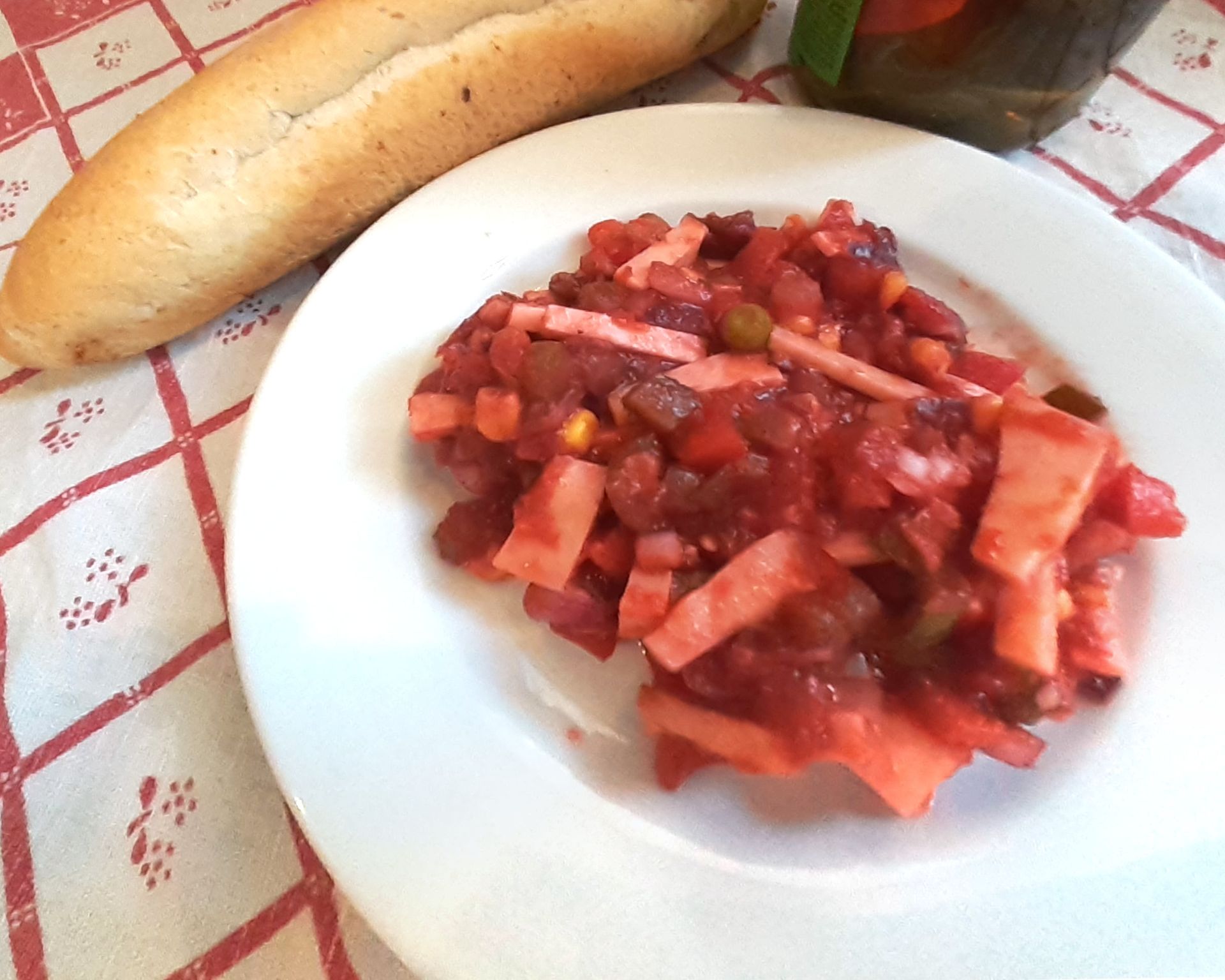 Červený šalát z feferóniek, párkov, hrášku, mrkvy, kukurice a uhoriek na bielom tanieri, v pozadí rožtek