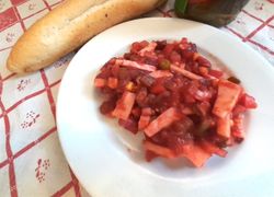 Červený šalát z feferóniek, párkov, hrášku, mrkvy, kukurice a uhoriek na bielom tanieri, v pozadí rožtek