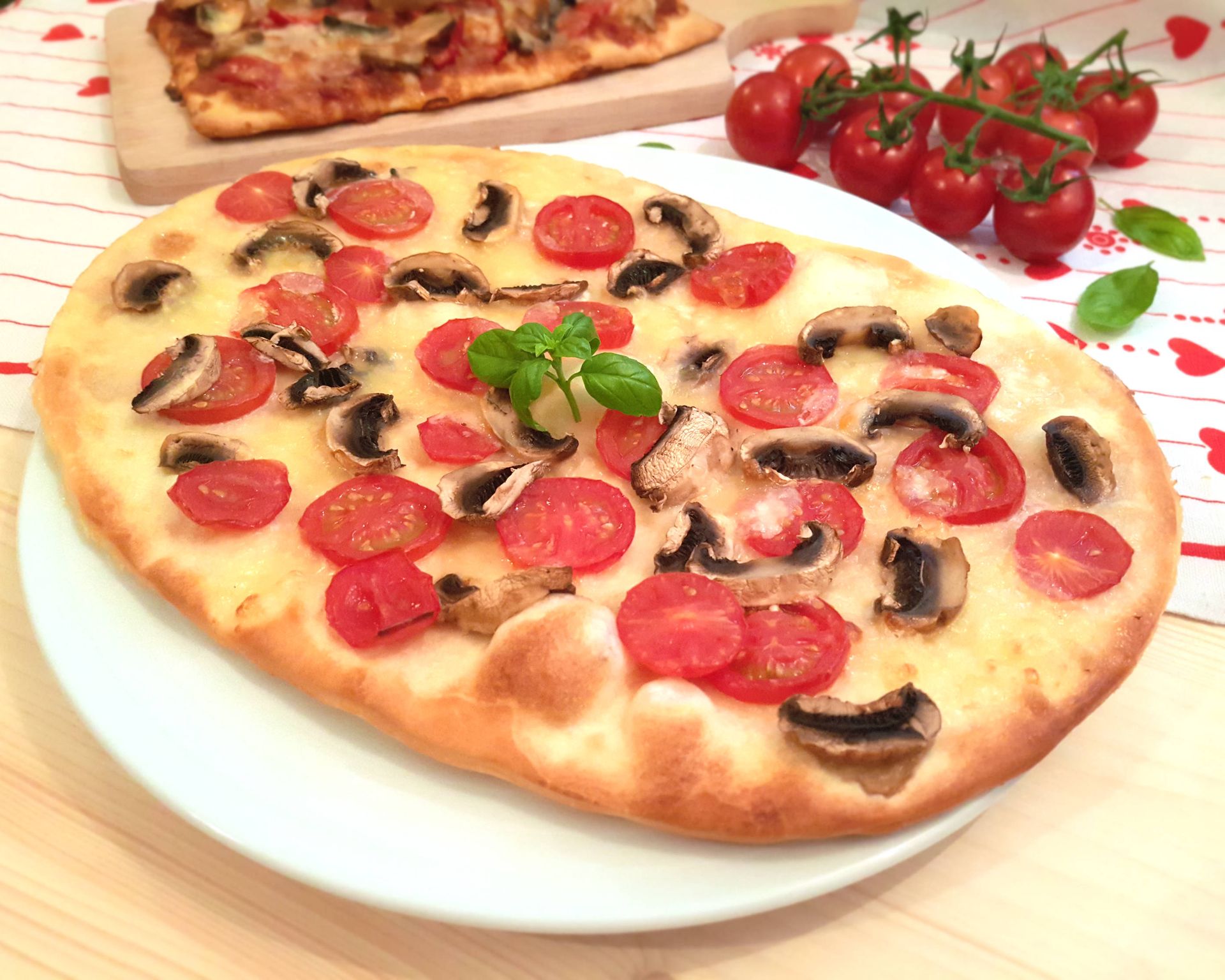 Smotanová pizza s mozzarellou, paradajkami a šampiňónmi na bielom pizza tanieri, v pozadí ďalšia pizza a cherry paradajky