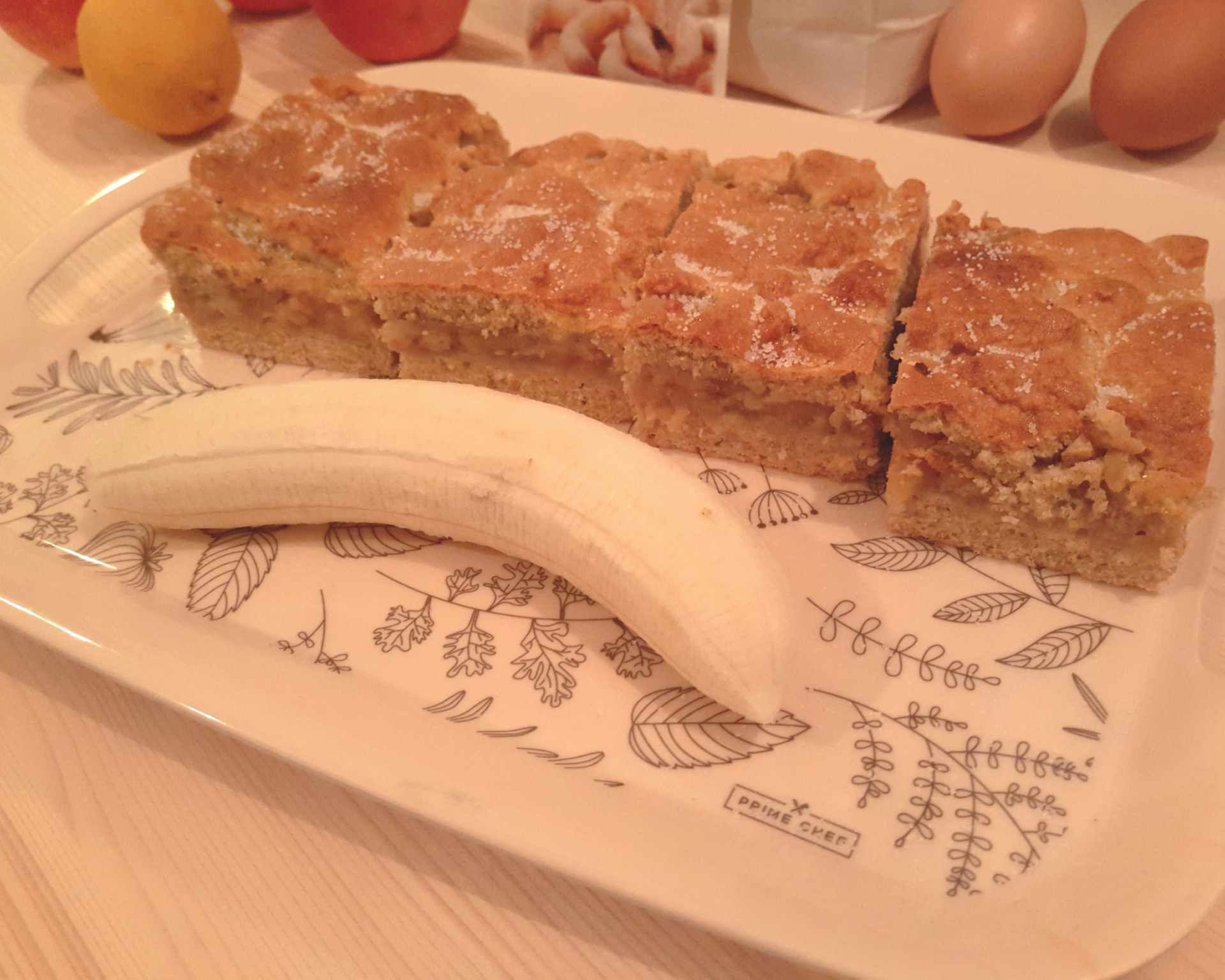 Jablkový koláč nakrájaný na obdĺžniky posypaný vanilkovým cukrom na tácke aj banán