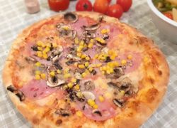 Šampiňónová pizza so šunkou, ktorá presahuje tanier, veľa kukurice, v pozadí sú paradajky.