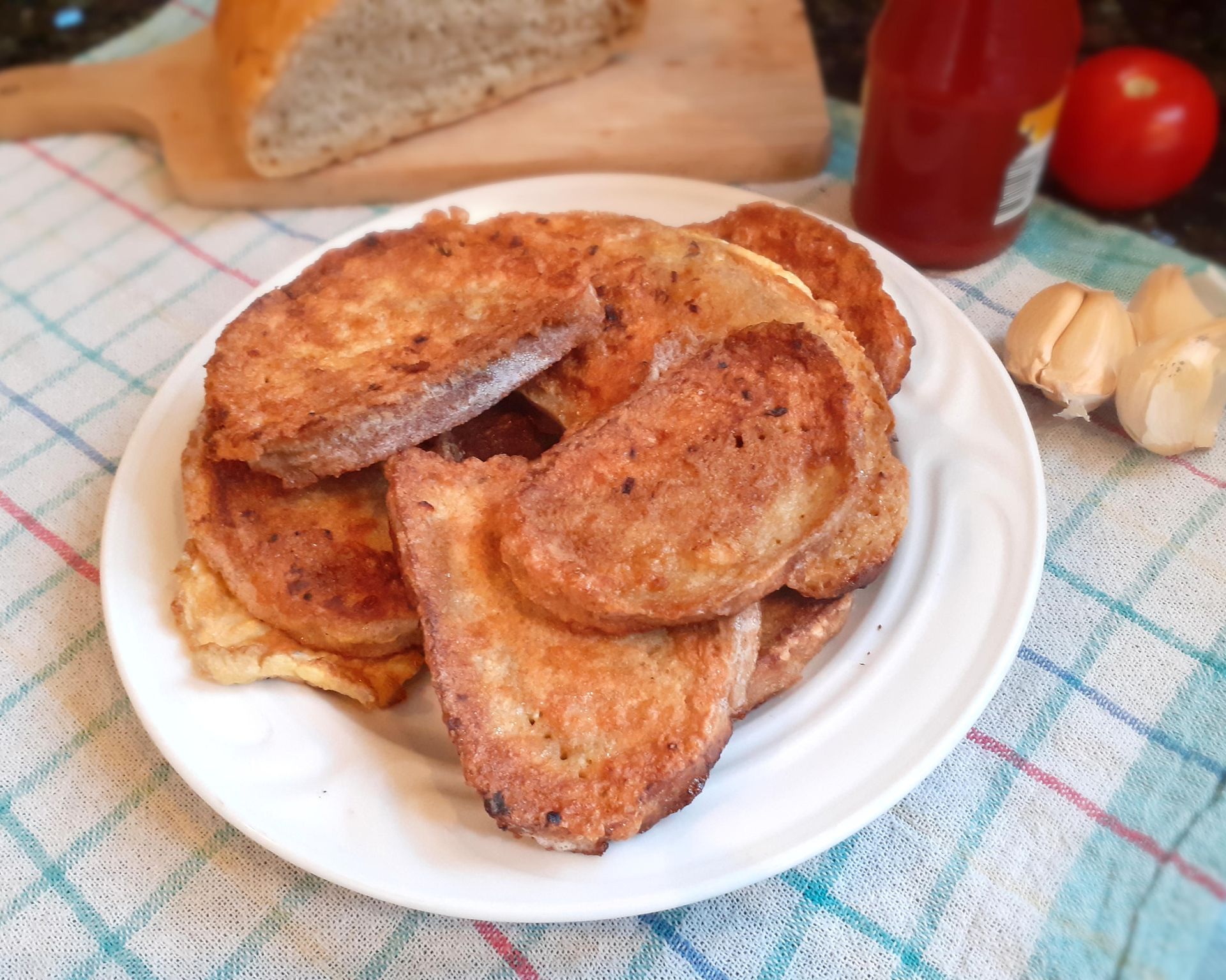 Suchý opečený chlebík vo vajci na bielom tanieri. V pozadí chlieb, kečup, paradajka a cesnak. Vajcový chlieb