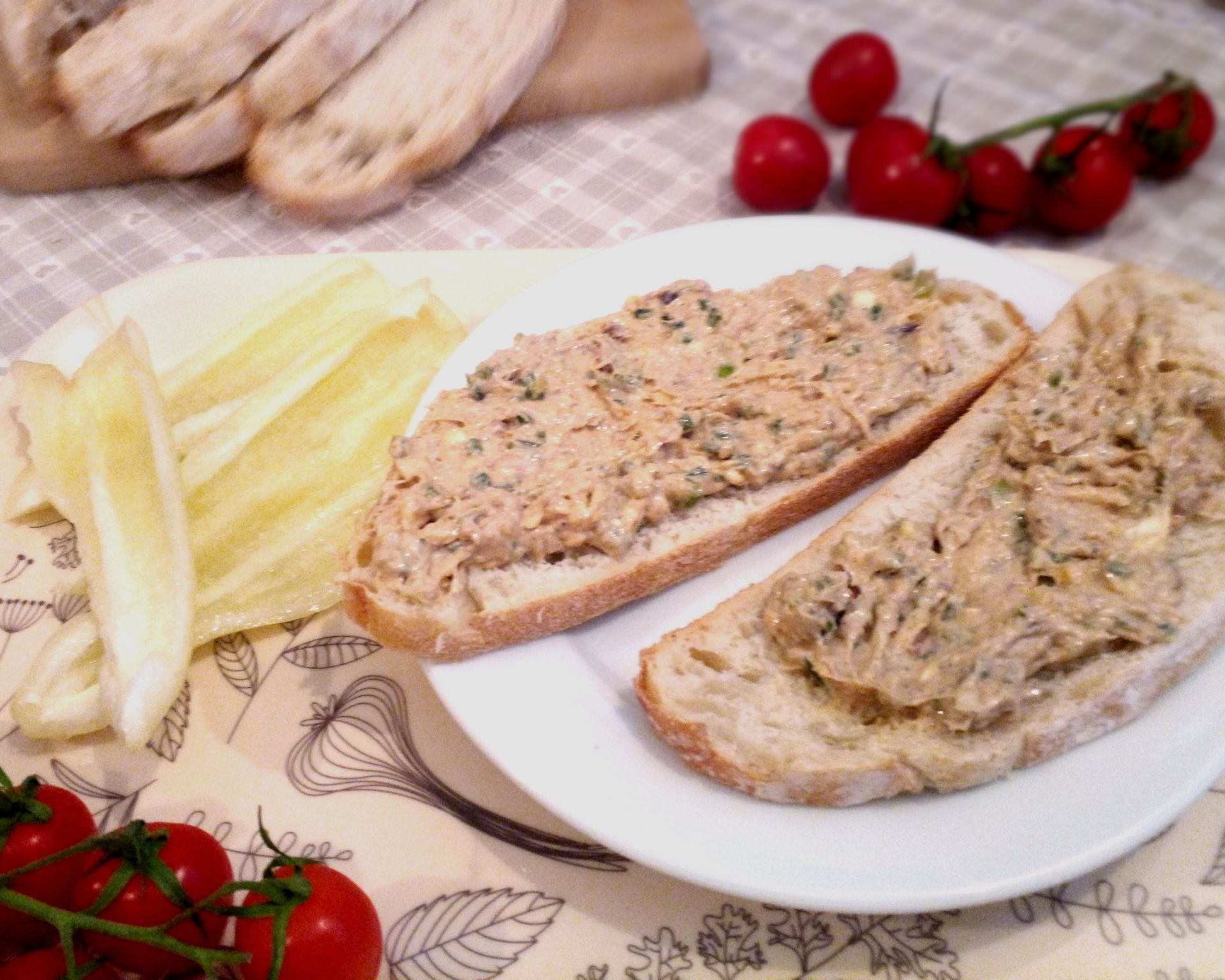 Dva krajce chleba natreté rybacou pomazánkou na tanieri, v pozadí nakrájaná paprika, cherry paradajky