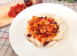 Penne s mexickou omáčkou, fazuľou, kukuricou a párkami v hlbokom tanieri, v pozadí cherry paradajky a chilli papričky