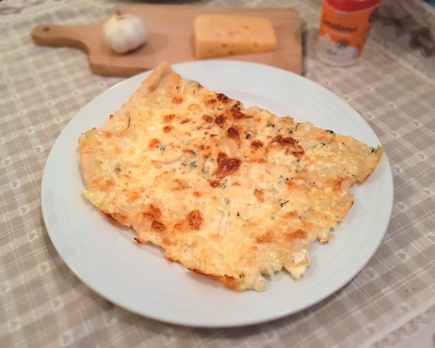 Kus smotanovo-syrovej pizze s Nivou na bielom tanieri, v pozadí drevená doska s cesnakom a ementálom