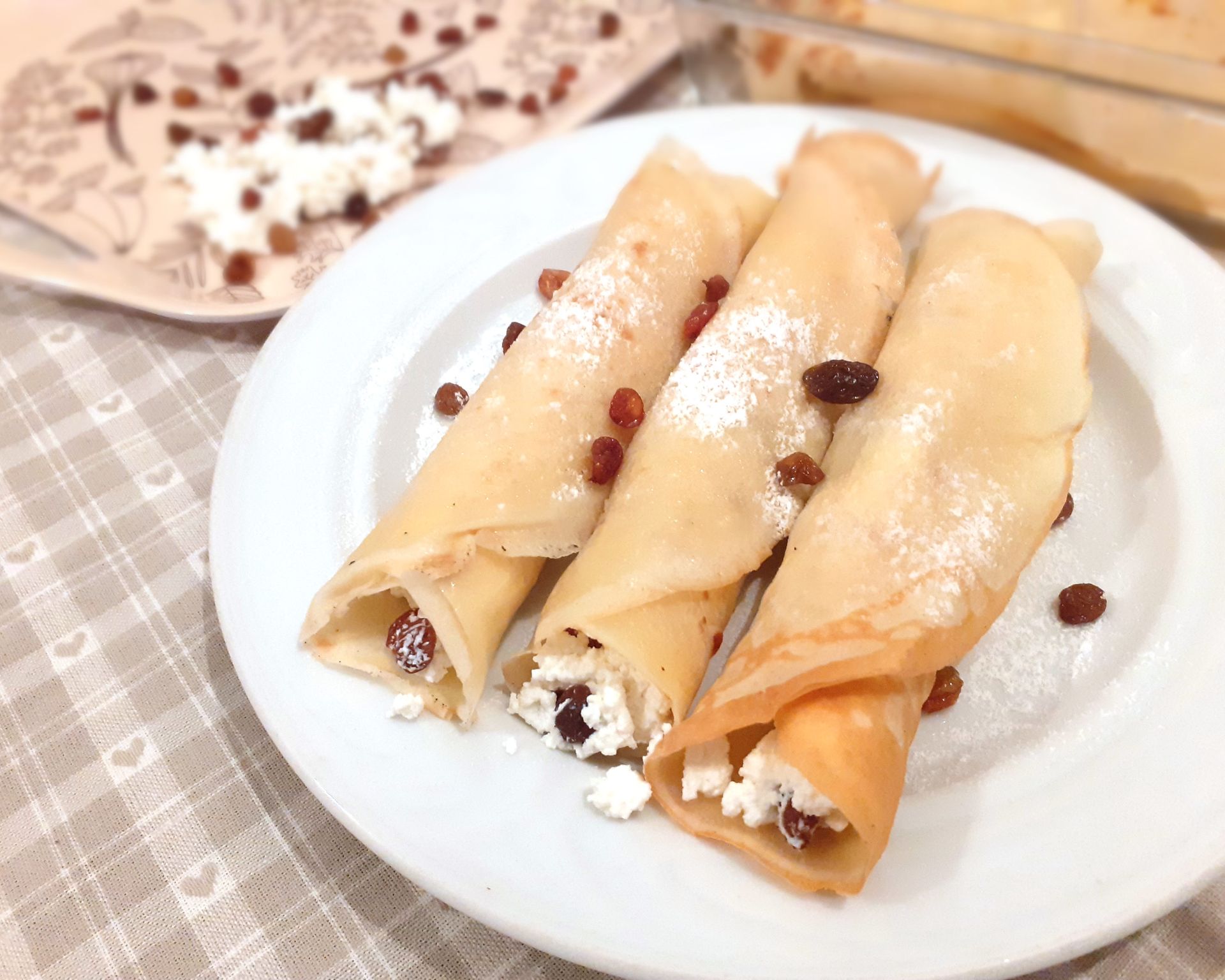 Tri zakrútené palacinky naplnené tvarohom, posypané práškovým cukrom, na vrchu hrozienka