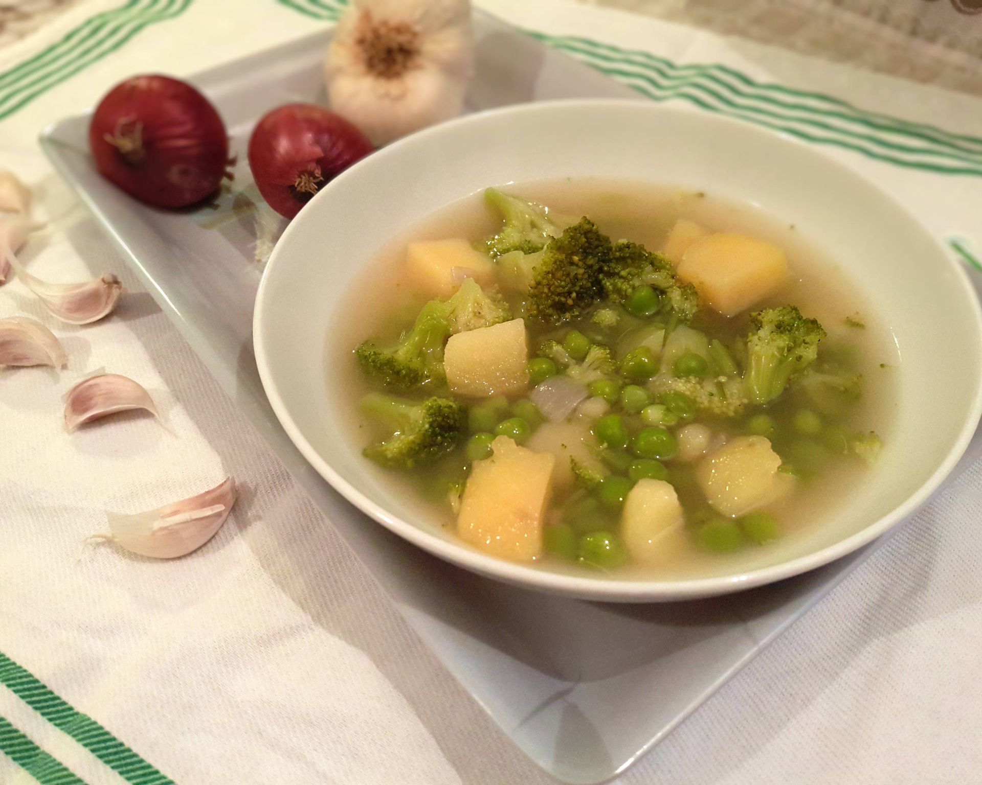 Tanier plný zelenej polievky s hrachom, brokolicou a zemiakmi, v pozadí cesnak, červená cibuľka