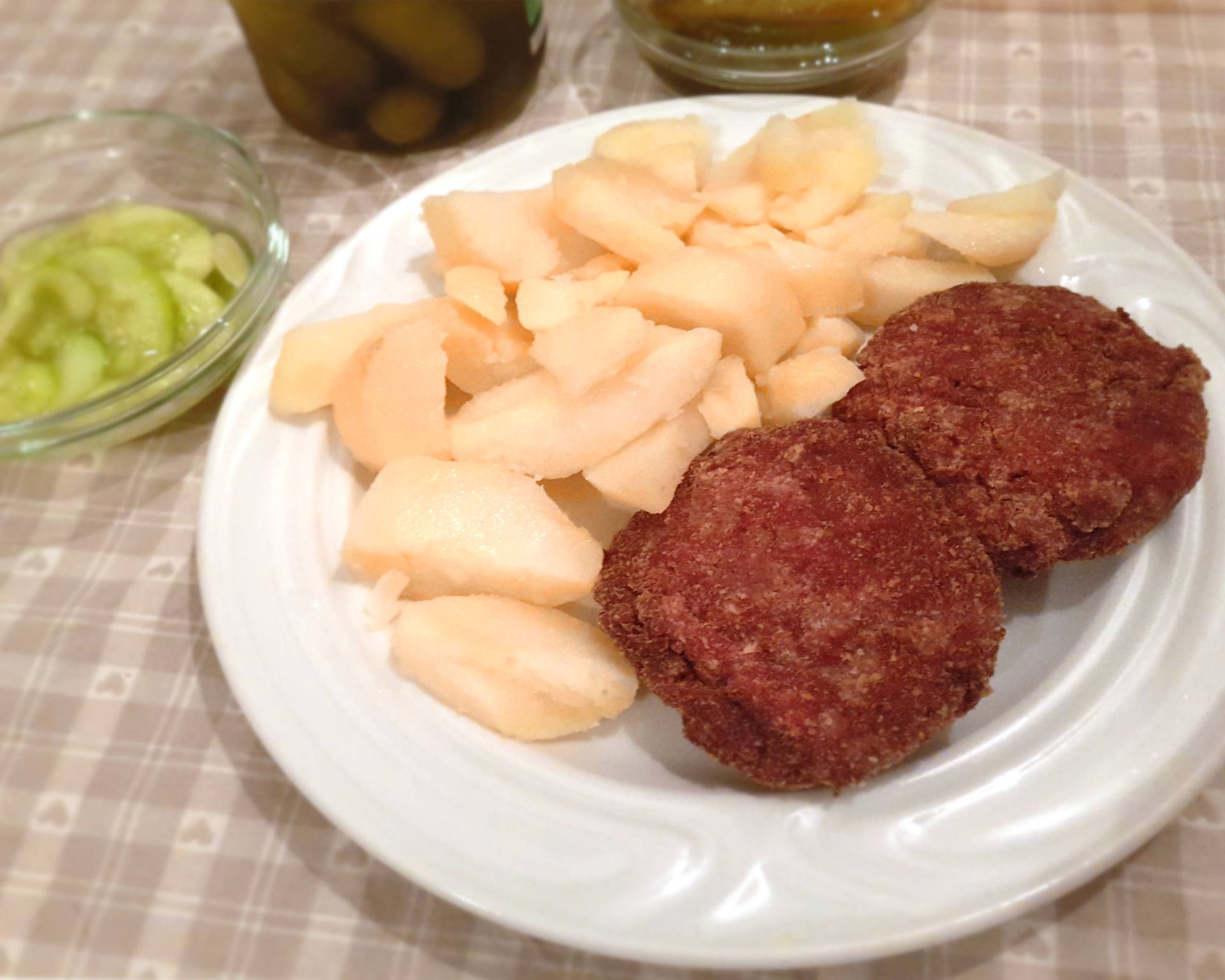 Karbonátky z mäsa plus varené zemiaky na bielom plytkom tanieri, v pozadí uhorkový šalát v miske a uhorky.