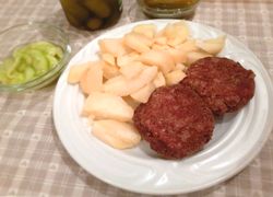 Karbonátky z mäsa plus varené zemiaky na bielom plytkom tanieri, v pozadí uhorkový šalát v miske a uhorky.